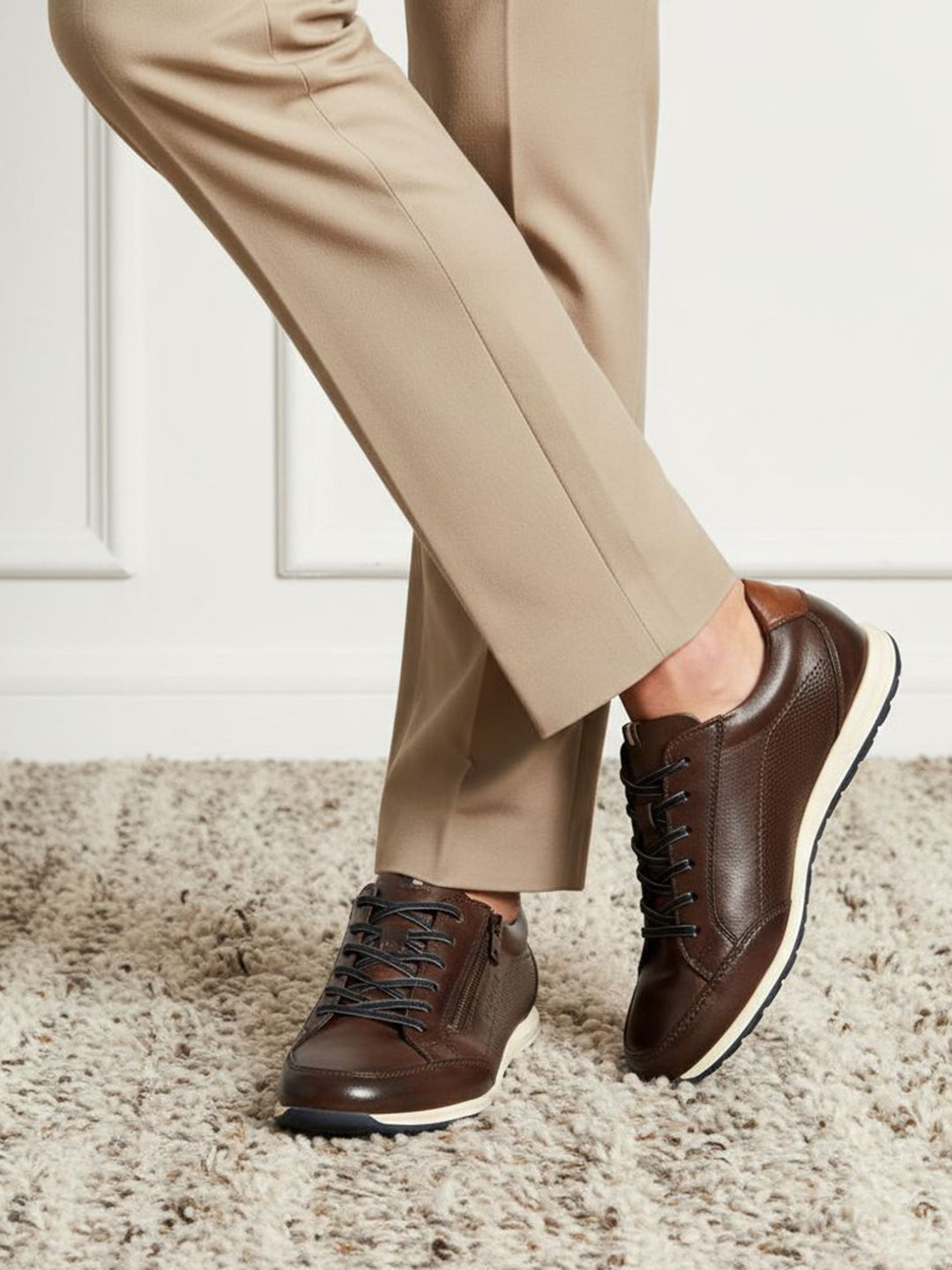 Lifestyle shot of bugatti Thorello brown premium leather sneakers with white soles, styled with tan trousers on a carpet.
