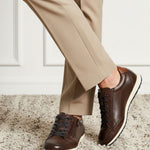 Lifestyle shot of bugatti Thorello brown premium leather sneakers with white soles, styled with tan trousers on a carpet.