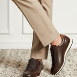 Lifestyle shot of bugatti Thorello brown premium leather sneakers with white soles, styled with tan trousers on a carpet.