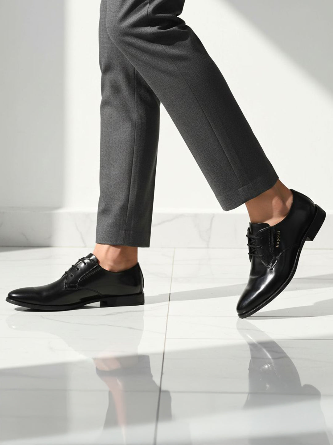 Side view of black bugatti leather derby shoes, paired with gray dress pants on a white marble floor.
