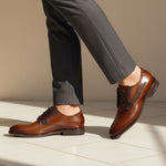 bugatti Sula cognac leather derby shoes, paired with gray dress pants, on a tiled surface.
