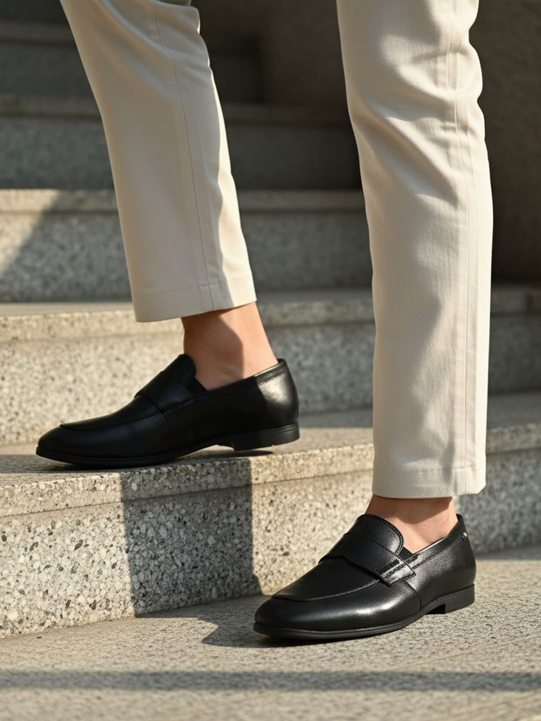 Black leather penny loafers by bugatti with a low heel, worn with light colored pants while standing on gray stairs.