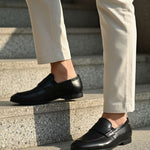 Black leather penny loafers by bugatti with a low heel, worn with light colored pants while standing on gray stairs.