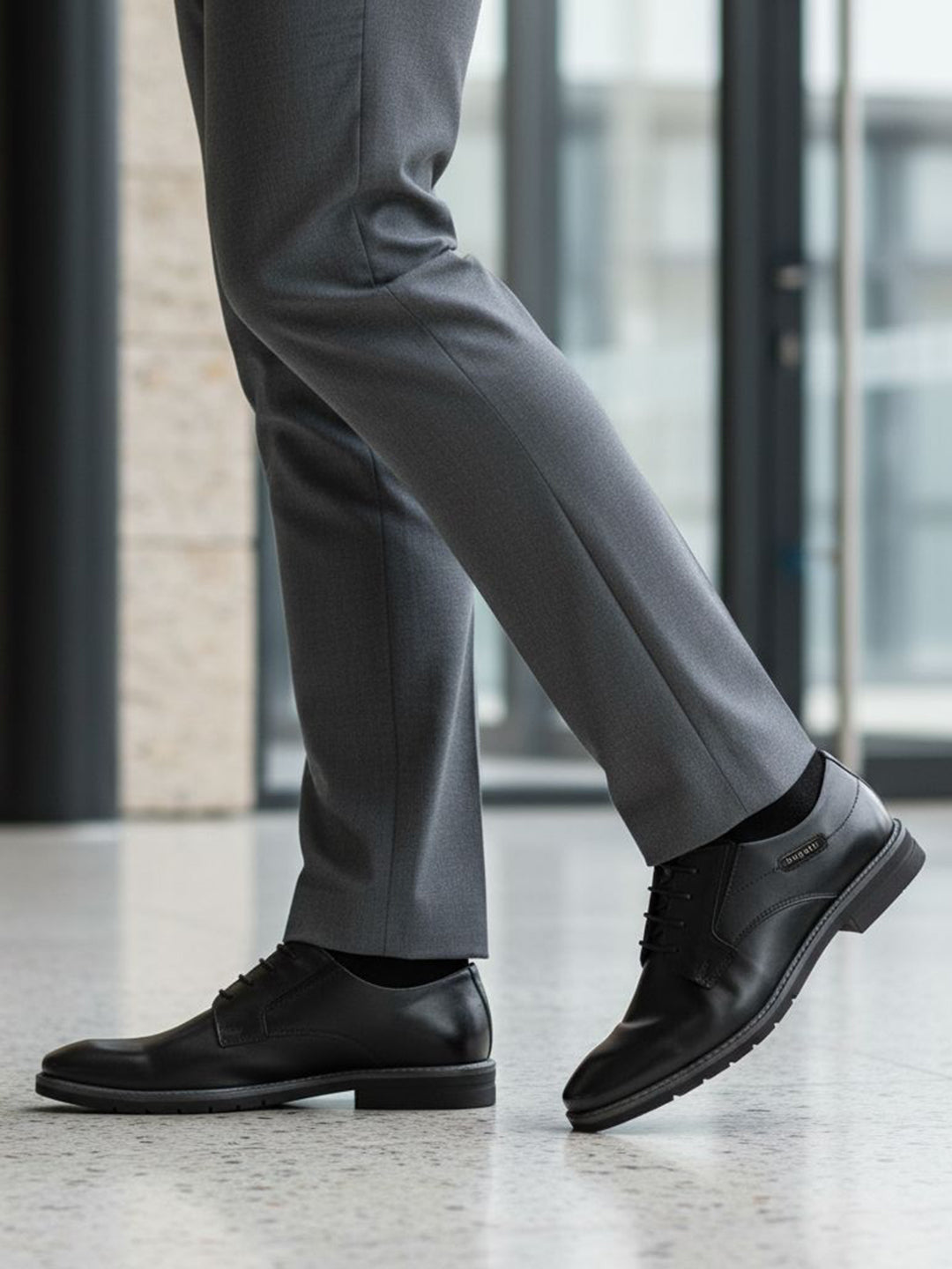 Lifestyle close-up of black bugatti Mero formal leather derby shoes, gray dress pants, and black socks.
