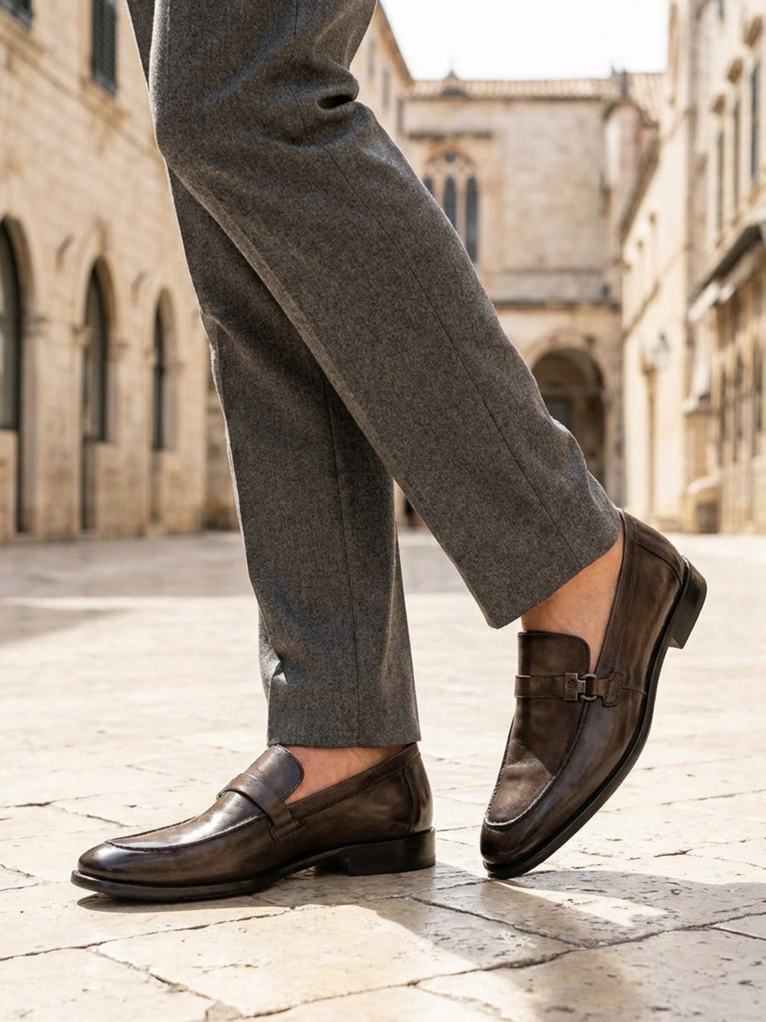 Lifestyle shot of bugatti dark green leather penny loafers with grey dress pants on a stone tiled street.

