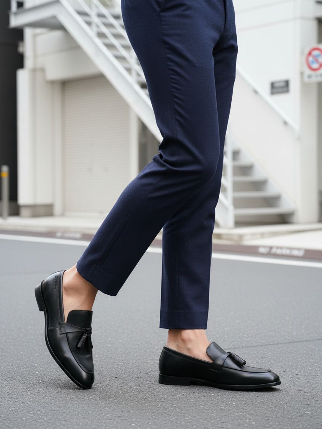 Cropped view of bugatti Rico black leather slip-on dress shoes with tassels, worn with navy trousers.
