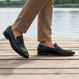 Lifestyle view of bugatti Rico black leather loafers with green trim, worn with tan pants on a wooden boardwalk.

