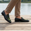 Lifestyle view of bugatti Rico black leather loafers with green trim, worn with tan pants on a wooden boardwalk.
