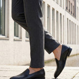 Cropped lifestyle shot of bugatti Rico black leather slip-on formal shoes, paired with gray dress pants.
