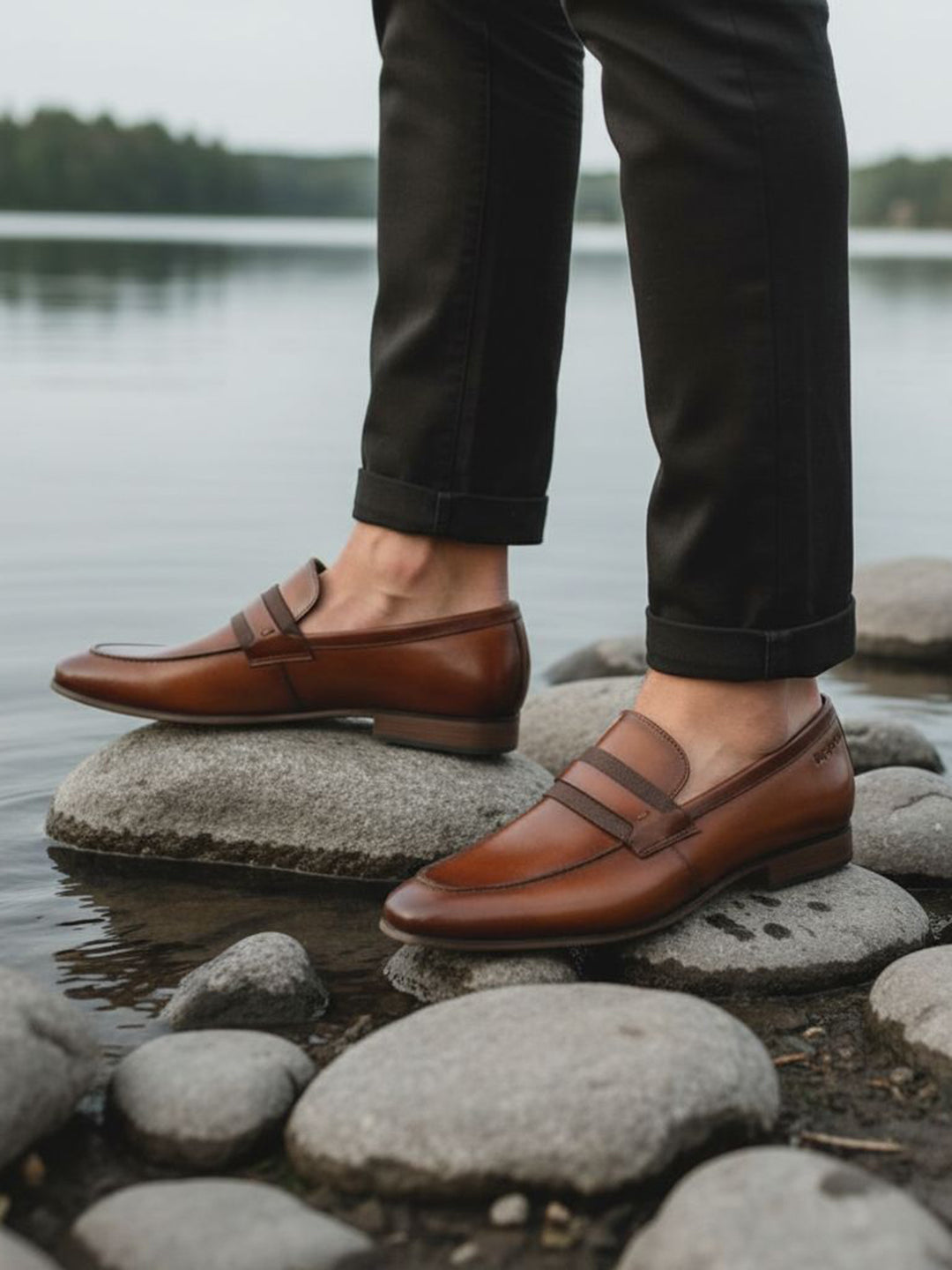 bugatti cognac leather loafers on rocks, paired with dark cuffed pants in a lifestyle shot.
