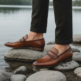 bugatti cognac leather loafers on rocks, paired with dark cuffed pants in a lifestyle shot.
