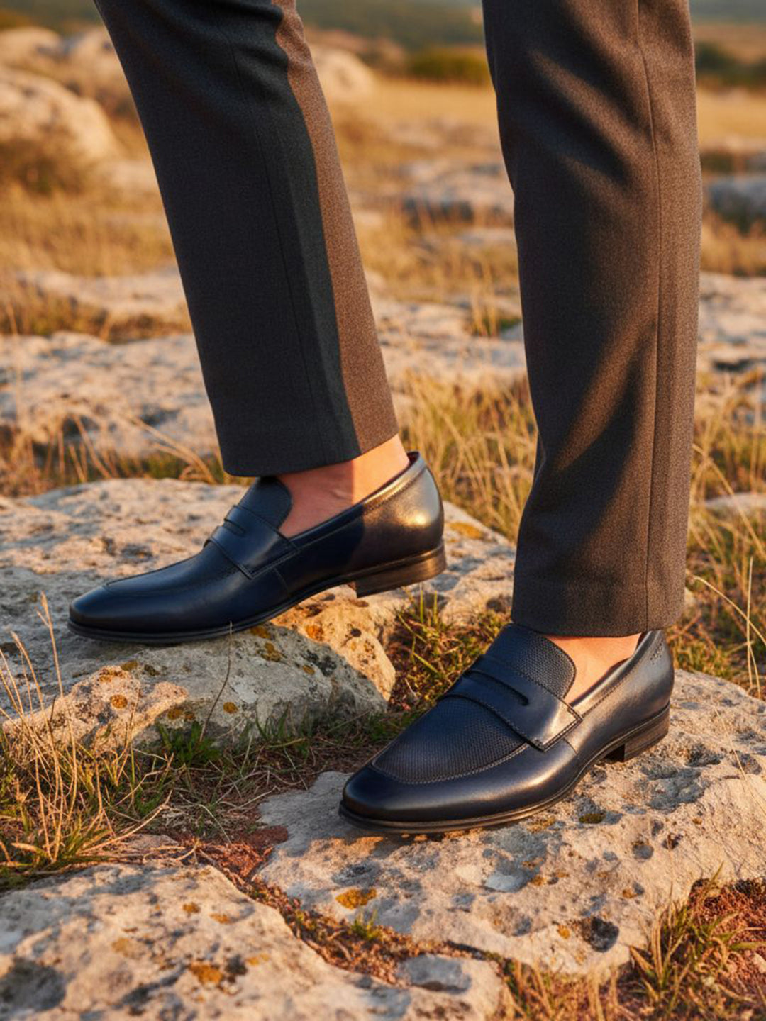 bugatti Margo dark blue premium leather penny loafers worn with gray dress pants on a rocky surface.
