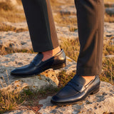 bugatti Margo dark blue premium leather penny loafers worn with gray dress pants on a rocky surface.
