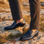bugatti Margo dark blue premium leather penny loafers worn with gray dress pants on a rocky surface.
