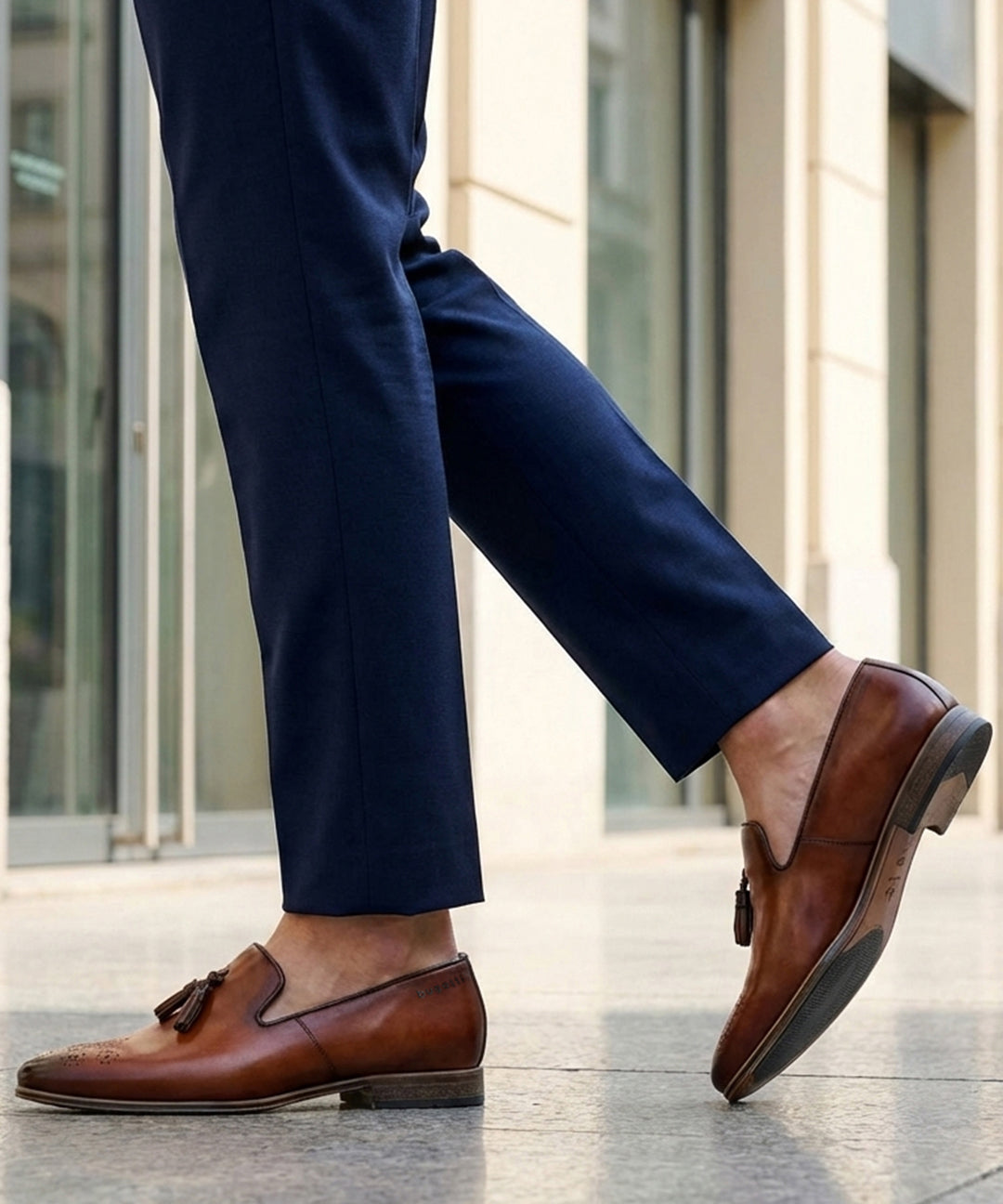 bugatti Margo cognac leather slip-on shoes with tassels, worn with dark blue pants in a lifestyle shot.
