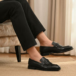 Lifestyle shot of black bugatti leather slip-on dress shoes, worn with black pants, and displayed on a rug.