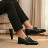 Lifestyle shot of black bugatti leather slip-on dress shoes, worn with black pants, and displayed on a rug.
