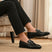 Lifestyle shot of black bugatti leather slip-on dress shoes, worn with black pants, and displayed on a rug.