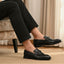 Lifestyle shot of black bugatti leather slip-on dress shoes, worn with black pants, and displayed on a rug.