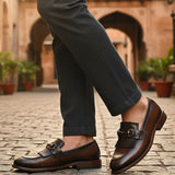 bugatti Liverta dark brown leather slip-on dress shoes with a decorative fringe, worn with gray pants.