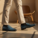 bugatti dark blue leather slip-on shoes with black socks and tan trousers, next to a wooden chair.
