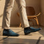 bugatti dark blue leather slip-on shoes with black socks and tan trousers, next to a wooden chair.
