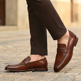 bugatti Mansueto cognac leather penny loafers with a textured strap detail, styled with brown trousers.
