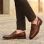 bugatti Mansueto cognac leather penny loafers with a textured strap detail, styled with brown trousers.
