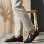 bugatti Tryst dark brown premium leather loafers with a classic penny strap, worn with beige pants.
