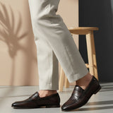 bugatti Tryst dark brown premium leather loafers with a classic penny strap, worn with beige pants.