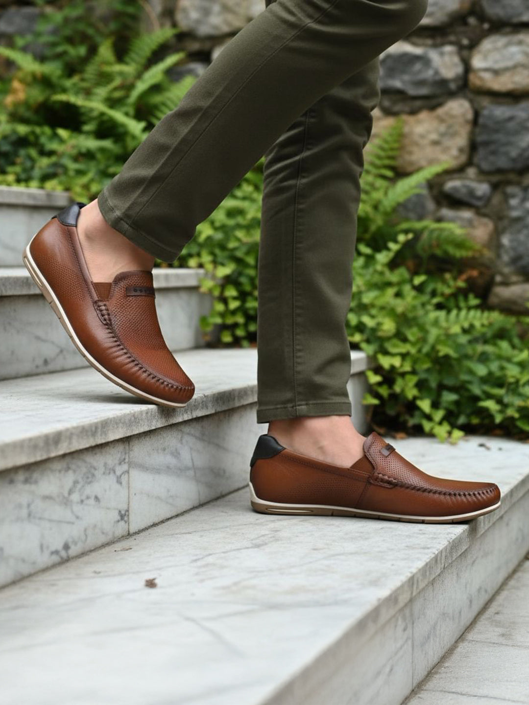 bugatti Chesley cognac leather slip-on shoe with a black accent at the heel, styled with olive pants on marble steps.
