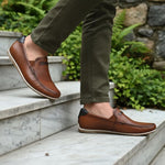 bugatti Chesley cognac leather slip-on shoe with a black accent at the heel, styled with olive pants on marble steps.
