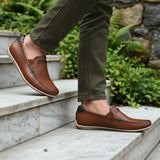 bugatti Chesley cognac leather slip-on shoe with a black accent at the heel, styled with olive pants on marble steps.
