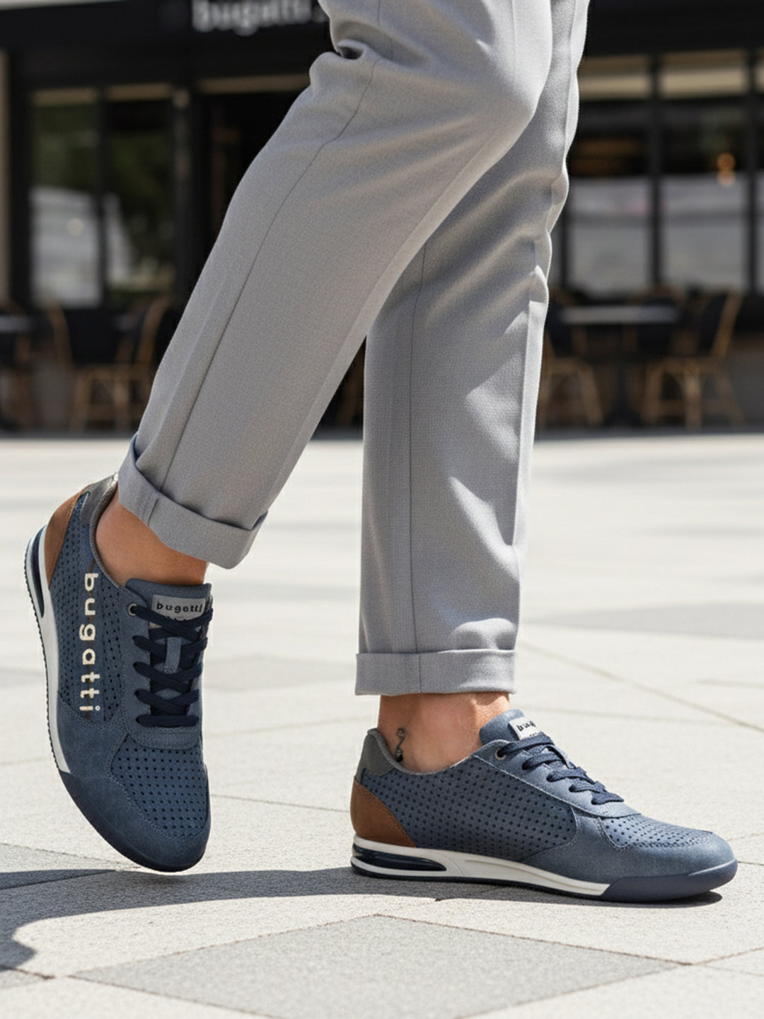 Lifestyle shot of bugatti Trevor blue Comfort Sneakers, a perforated leather shoe with brown heel detail, worn with gray slacks.
