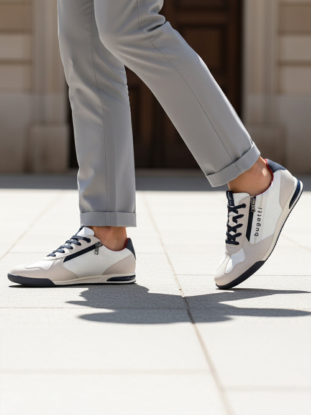 Lifestyle shot of bugatti Trevor white sneakers, with zipper detail, worn with gray pants and walking on pavement.