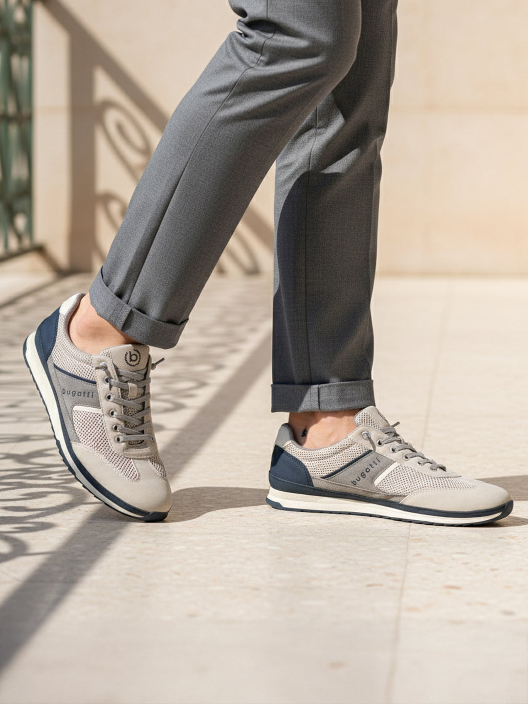 Lifestyle image of bugatti Riptide sand Comfort Sneakers, with gray mesh and suede upper, dark blue accents, paired with gray trousers.
