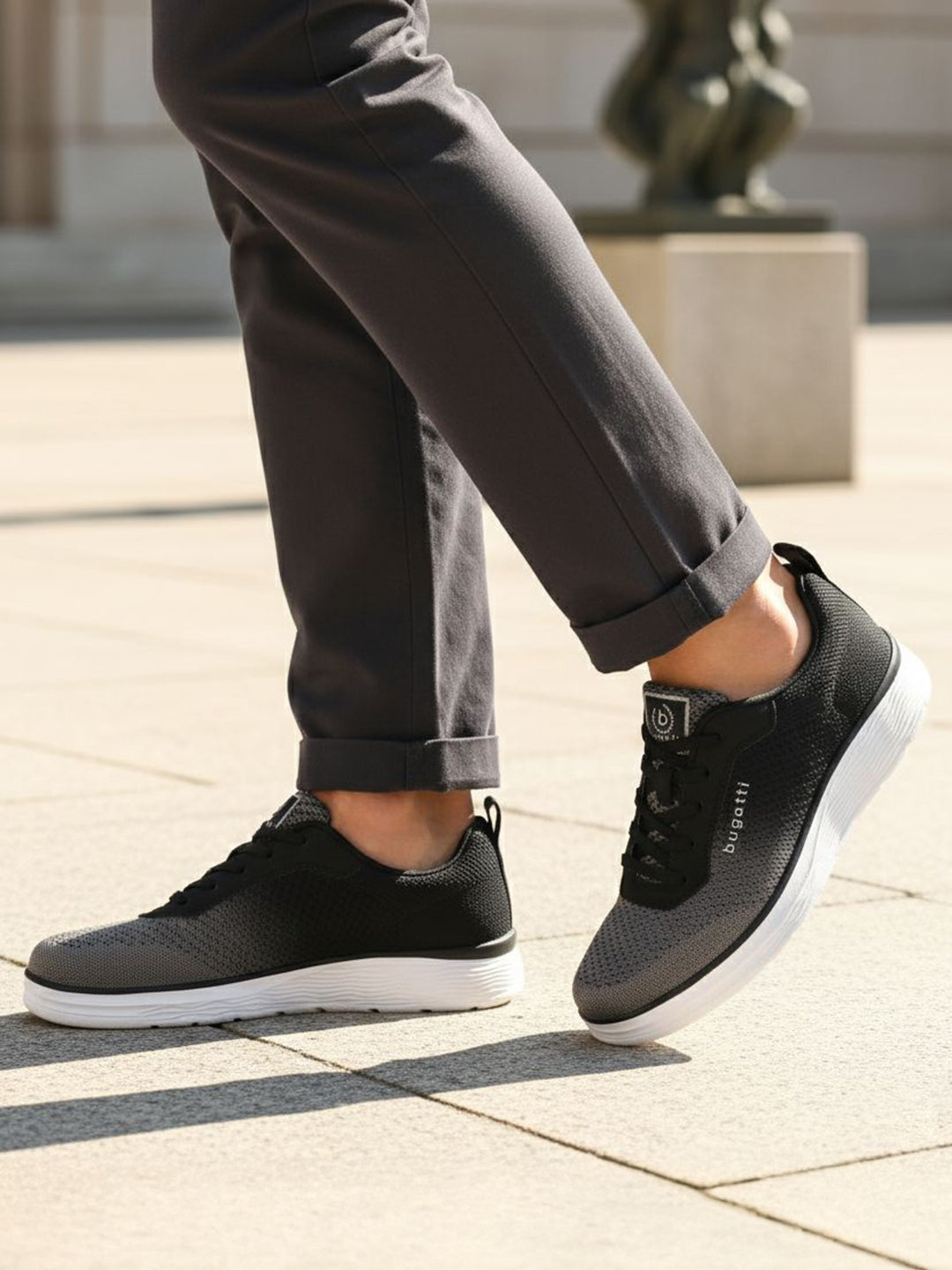 Lifestyle image of bugatti Astra athleisure sneakers in dark grey with a white sole, worn with rolled grey pants.

