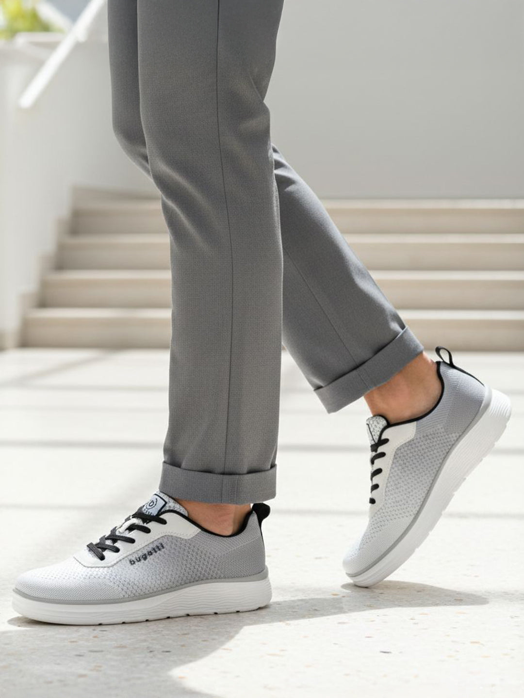 Lifestyle shot of bugatti Astra athleisure sneakers in white with black laces, paired with grey pants.
