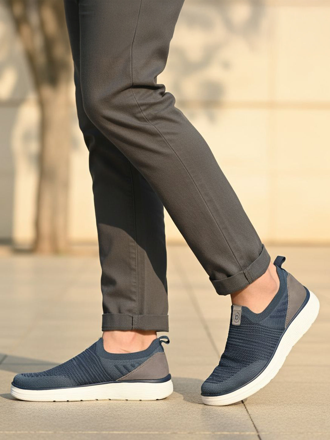 bugatti Astra blue slip-on casual shoes with a knit texture and white soles, styled with gray trousers.
