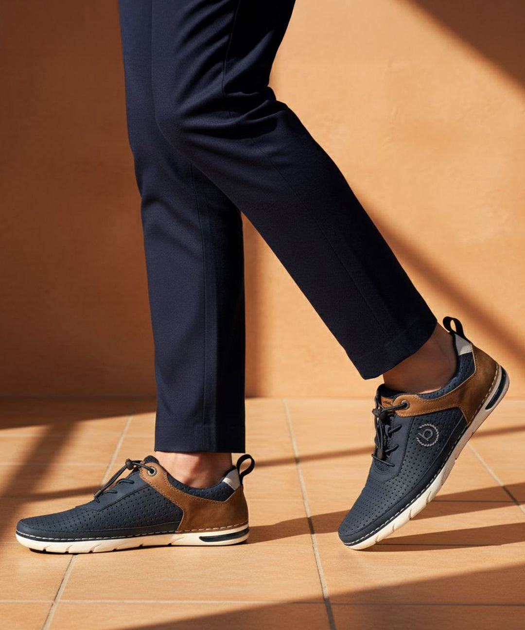 Lifestyle close-up of bugatti Glenans dark blue comfort sneakers with brown accents and white soles, paired with navy dress pants.