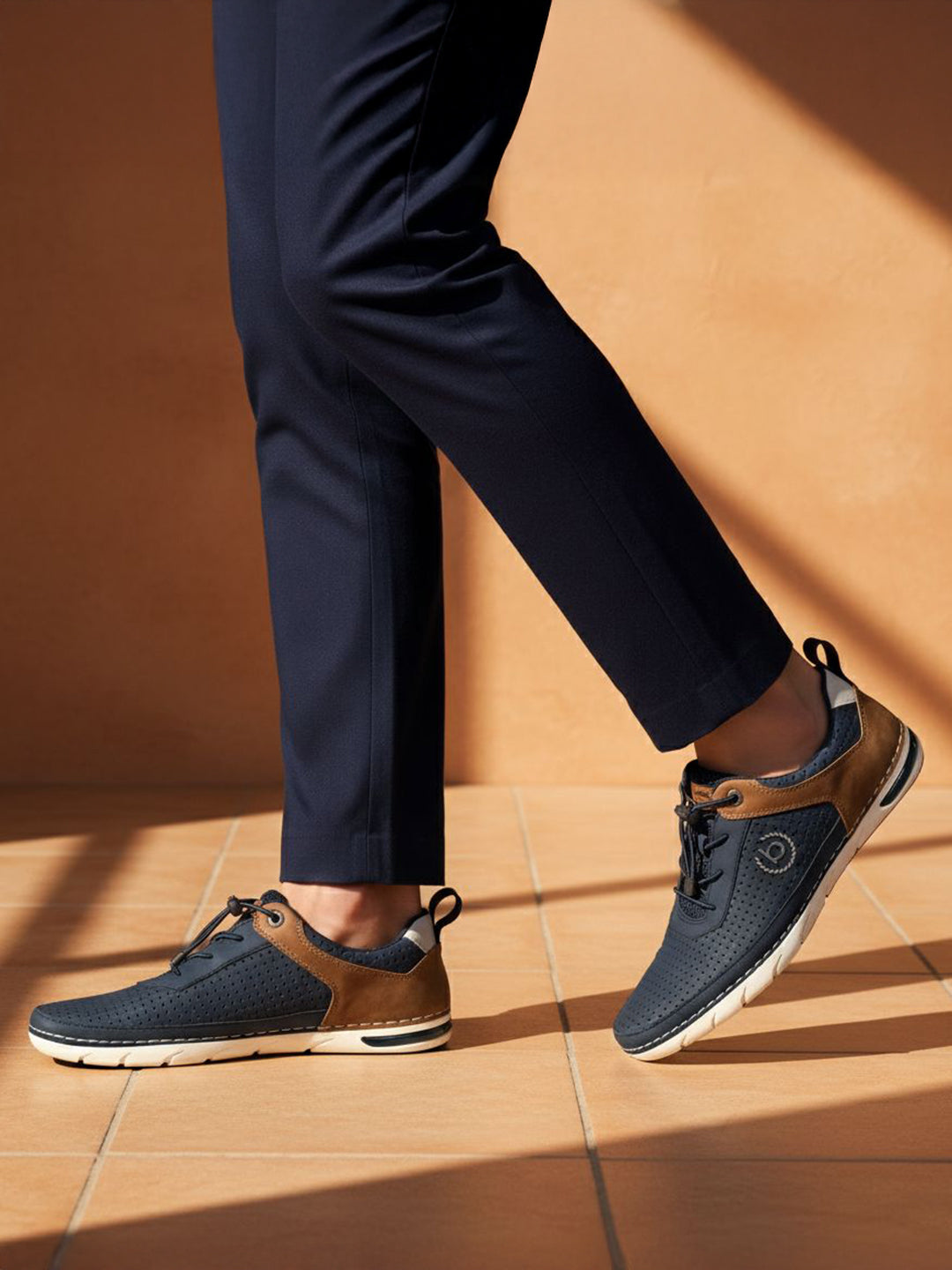 Lifestyle close-up of bugatti Glenans dark blue comfort sneakers with brown accents and white soles, paired with navy dress pants.