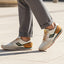 Lifestyle image of bugatti Sacramento offwhite comfort sneakers with green accents, brown heel, paired with gray dress pants.