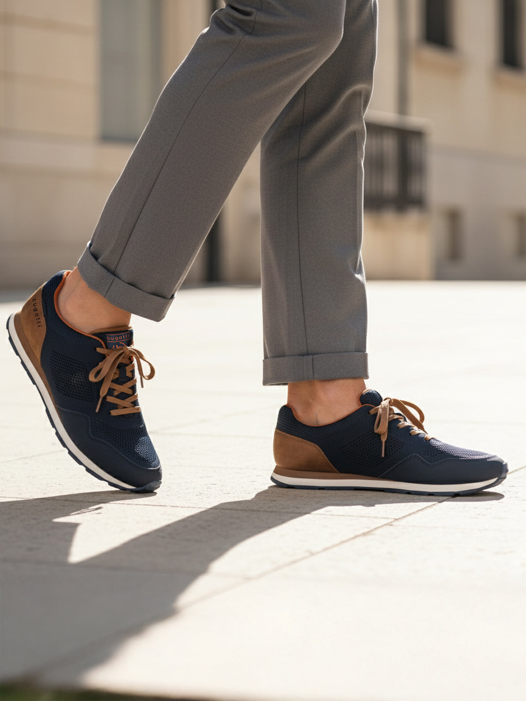 Lifestyle image of bugatti Sacramento dark blue sneakers with brown accents and laces, worn with cuffed grey trousers.
