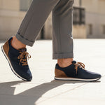 Lifestyle image of bugatti Sacramento dark blue sneakers with brown accents and laces, worn with cuffed grey trousers.
