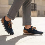 Lifestyle image of bugatti Sacramento dark blue sneakers with brown accents and laces, worn with cuffed grey trousers.

