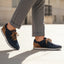 Lifestyle image of bugatti Sacramento dark blue sneakers with brown accents and laces, worn with cuffed grey trousers.
