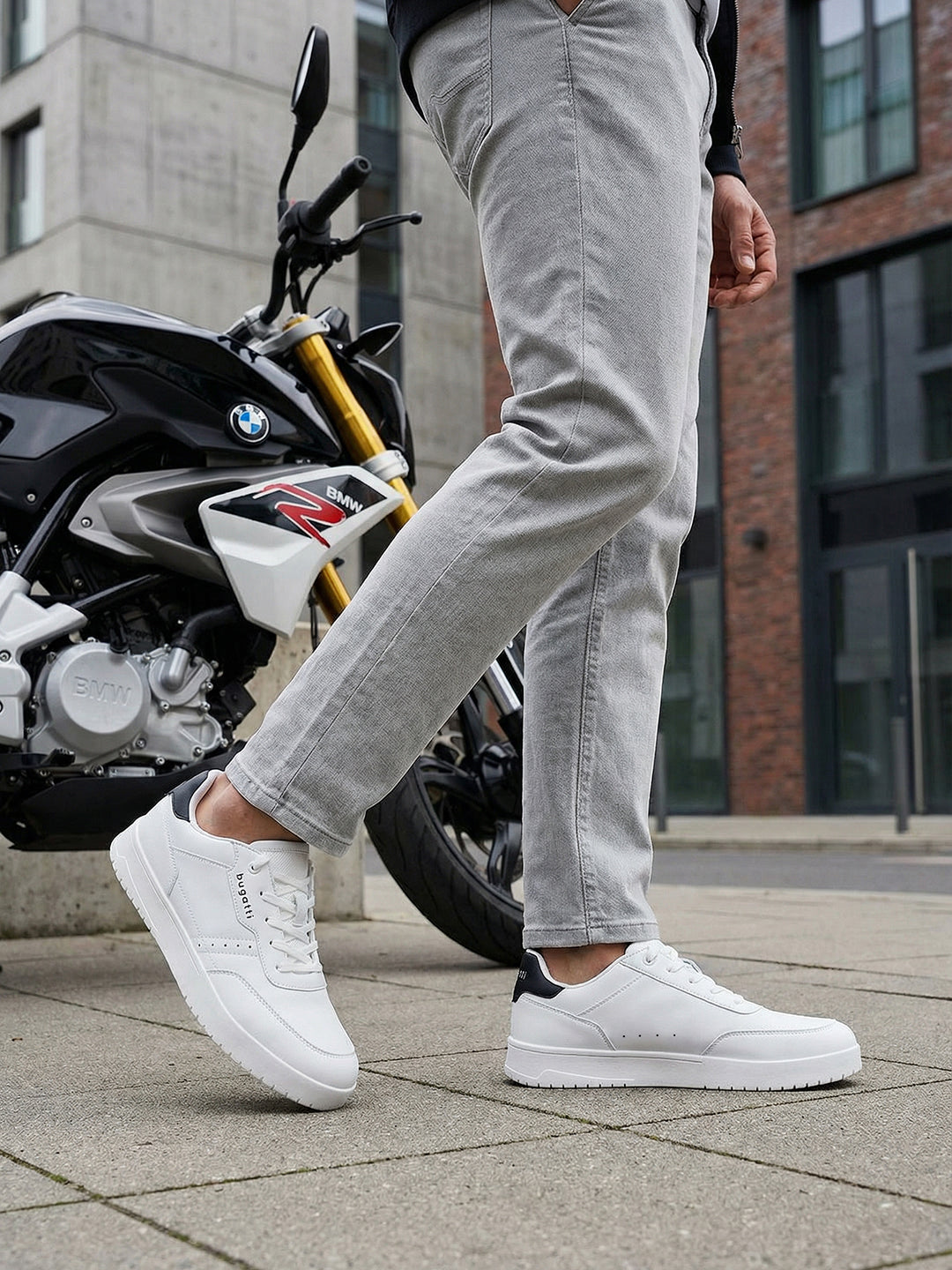 Lifestyle close-up of white bugatti Helium Comfort Sneakers, worn with grey pants and standing next to a motorcycle.
