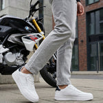 Lifestyle close-up of white bugatti Helium Comfort Sneakers, worn with grey pants and standing next to a motorcycle.
