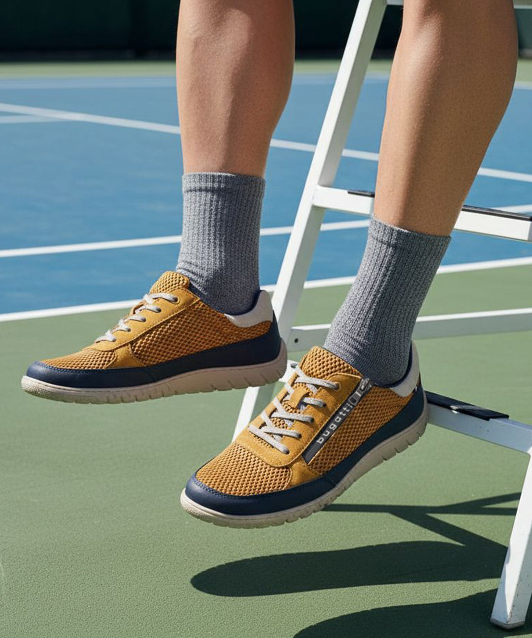 Lifestyle shot of bugatti Bermudas yellow comfort sneakers worn with gray socks on a tennis court.
