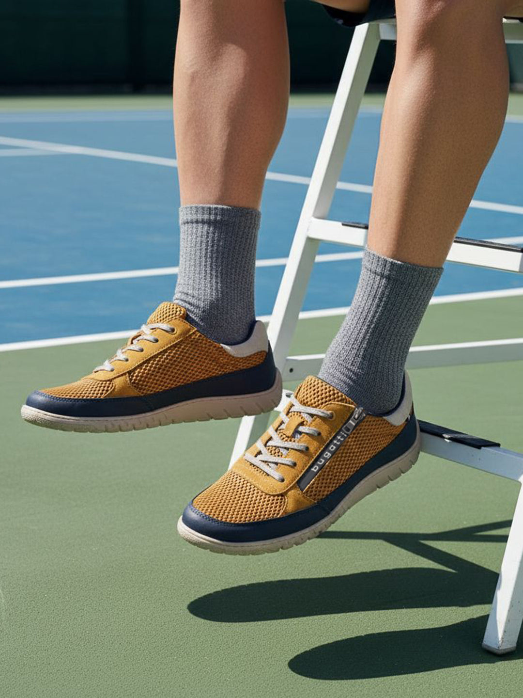Lifestyle shot of bugatti Bermudas yellow comfort sneakers worn with gray socks on a tennis court.

